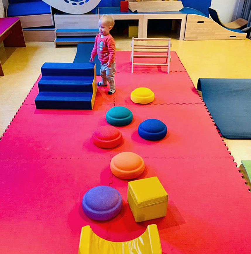 Kleinkind steht im Spielraum mit bunten Balance-Steinen auf roter Matte